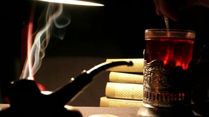 夜间学习室内: 一个人在玻璃杯里的一杯茶中混合糖，烟斗冒烟，一堆书躺在灯光下