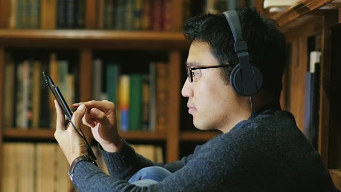 一个戴着眼镜的亚洲年轻人喜欢平板电脑。坐在书架背景上的图书馆