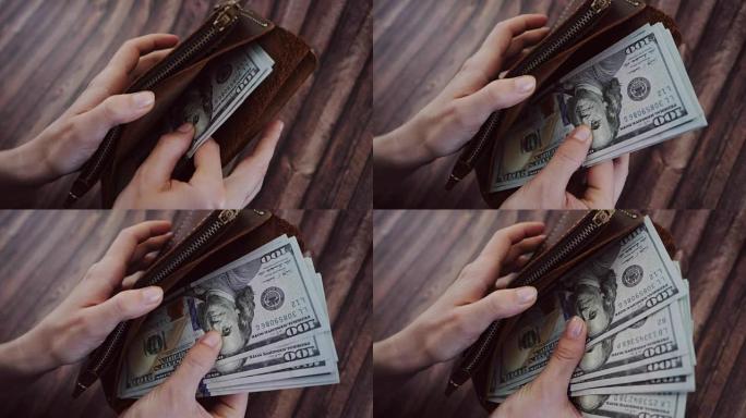 一个女人数数并从她的棕色皮革钱包中取出美元的俯视图。女性在木制背景下拿着钱包。慢动作