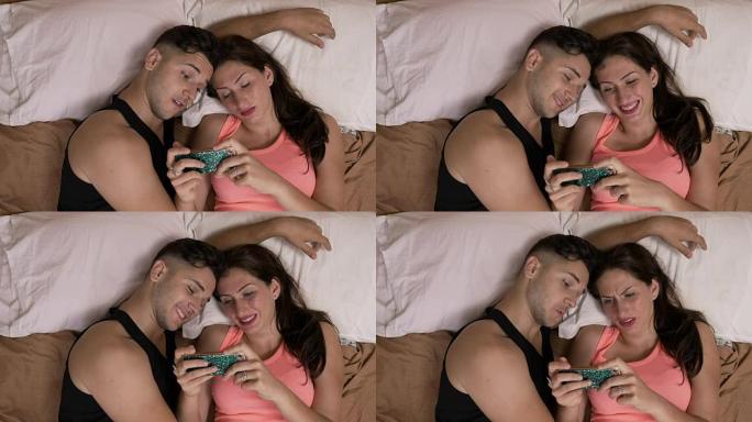 娱乐的年轻夫妇坐在床上看智能手机上的视频，表达了他们对内容的惊讶反应
