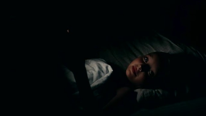 一个女孩躺在床上看电话里的恐怖。夜间电影观看。白人女孩晚上不睡觉。卧室里的女孩看视频，害怕做噩梦。