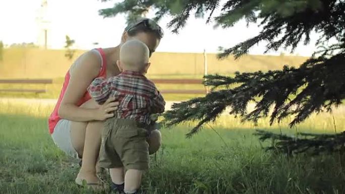 母亲站在公园里靠近树木，抱着微笑的小男孩儿子。儿童触摸树