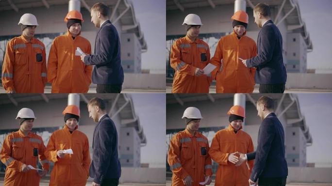 穿着西装的经理给穿着橙色制服和头盔的工人信封。项目的成功完成。慢动作