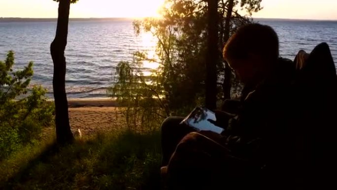 少年少年坐在大湖岸边的椅子上。十几岁的男孩热情地在平板电脑上玩电脑游戏