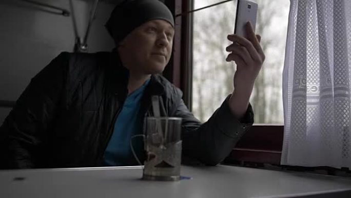 一位年轻的英俊男子坐火车并进行视频通话，微笑，比划手势，在窗外用玻璃特制杯子喝茶自然背景