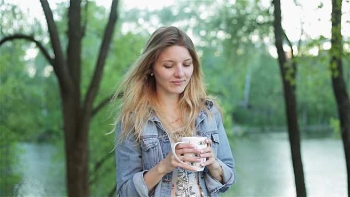 长发快乐的女孩在模糊的自然背景下手里拿着一杯茶或咖啡。穿着牛仔裤夹克的漂亮女人微笑着喝饮料。慢动作镜
