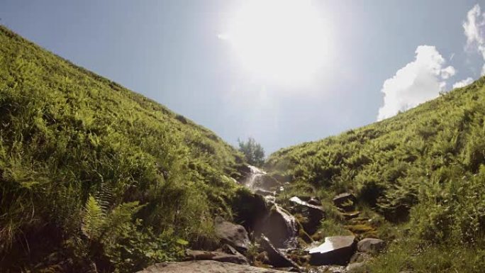 溪流在两个绿色的山丘之间流动，阳光照在相机的蓝天中