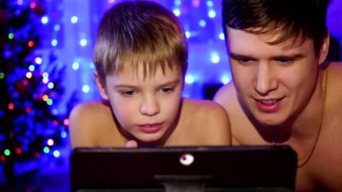 孩子和父亲看着躺在床上的平板电脑。背景灯花环