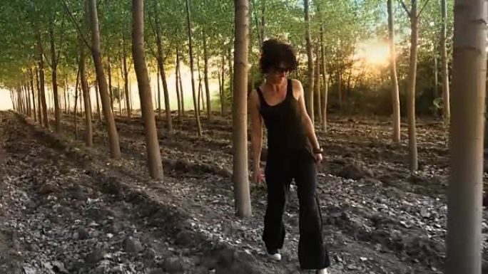 日落时，美丽的黑发女人走在森林树丛中，阳光照在树干后面