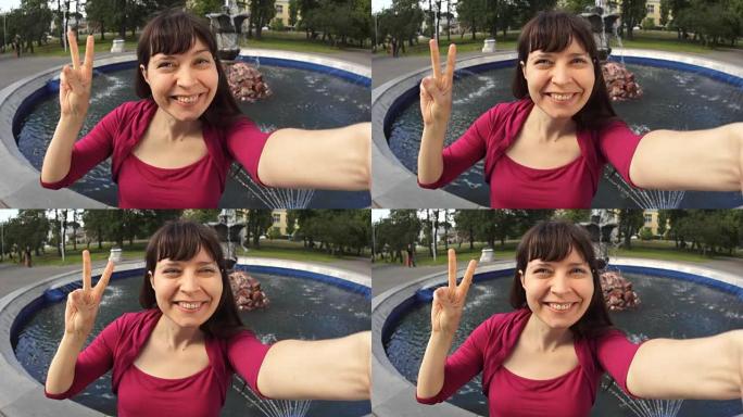 显示胜利标志的年轻女子坐在公园的喷泉旁自拍