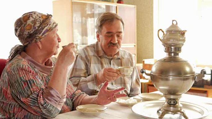 一对老年夫妇正在用老式的俄罗斯水壶茶炊喝茶。一个留着小胡子的男人和妻子穿着方巾说话