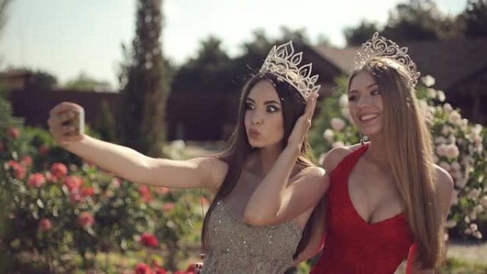 两个穿着晚礼服和皇冠的女孩在花园里用玫瑰在手机上自拍