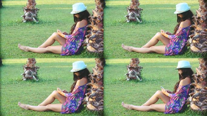 夏天，年轻的晒黑的长发女人坐在热带棕榈树下。她使用智能手机