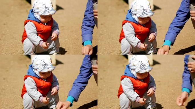 一岁的孩子在操场上玩沙子。母亲附近帮助他玩游戏。穿着红色马甲和白色头饰的男孩