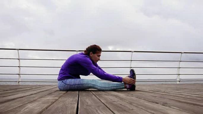 运动女子清晨坐在海边的平台上伸展双腿。夏天在海滩上训练