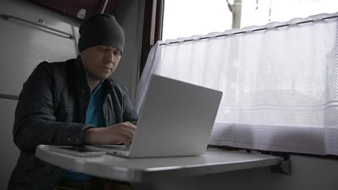 货车火车，一个男人坐在桌子旁，输入灰色的笔记本电脑，推开窗帘，看着窗外。窗外，树木生长