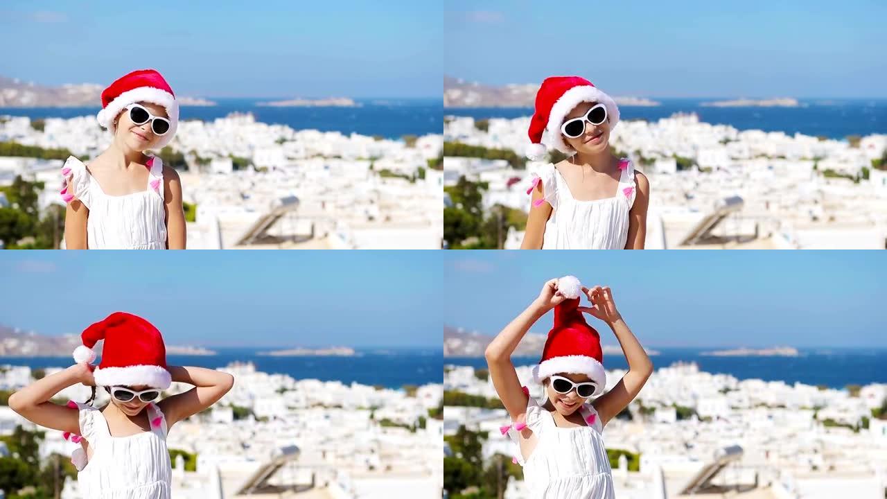 小女孩在圣诞老人帽子户外背景米克诺斯老街。孩子在希腊米科诺斯岛上典型的希腊传统村庄的街道上，白色的墙