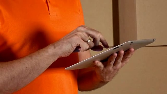 穿着橙色制服的仓库工人在棕色纸箱背景下使用平板电脑。全高清特写视频