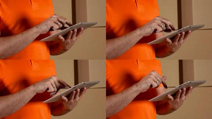 穿着橙色制服的仓库工人在棕色纸箱背景下使用平板电脑。全高清特写视频