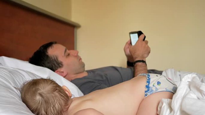 父亲在熟睡的婴儿附近的平板电脑上阅读新闻。一个男孩在床上的房子的前景中休息
