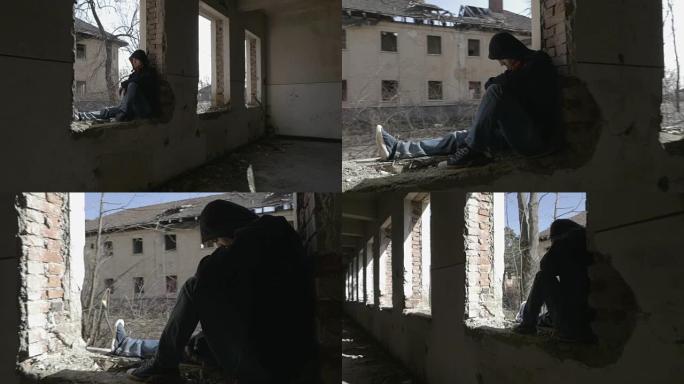 蒙面沮丧的年轻人坐在一栋废弃建筑的窗框上