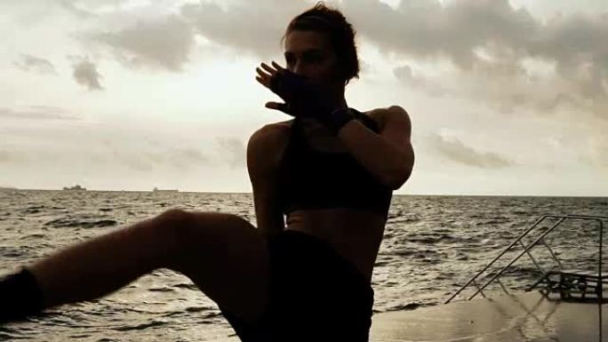 女拳击手使她的腿对着太阳的海边在慢动作侧踢。美丽的女拳击手早上在沙滩上训练，拳打脚踢