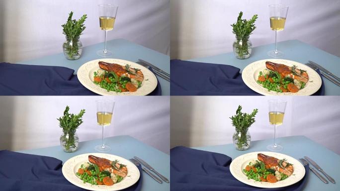 在一张有蓝色桌布的桌子上是一个盘子，上面放着烤猪肉和酱汁和蔬菜装饰，后面放着一个白葡萄酒缸和一个装有