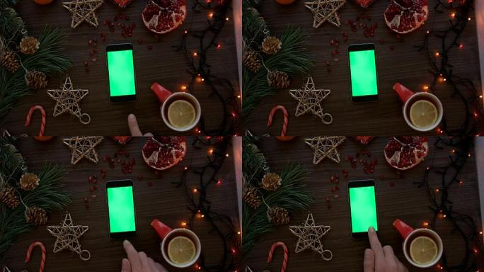 顶视图手使用绿屏智能手机。触摸屏上的手指滚动页面。木制桌子背景上的圣诞装饰。色度键