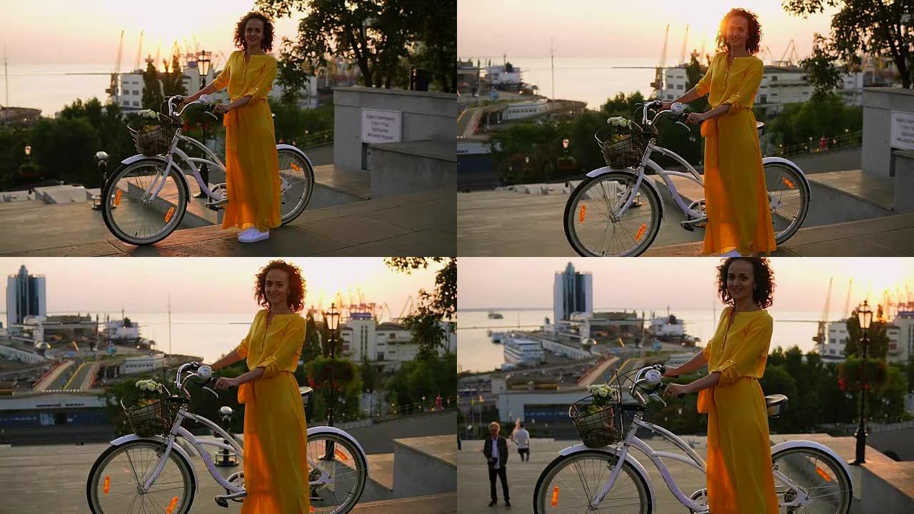 清晨，穿着黄色长裙的微笑黑发女人站在她的城市自行车旁边，拿着车把，篮子里放着鲜花。镜头耀斑，美丽的城