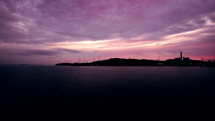 在暴风雨的海洋海岸线上，发电站和风力发电机的轮廓在日出或日落时带有辐射的火焰云。