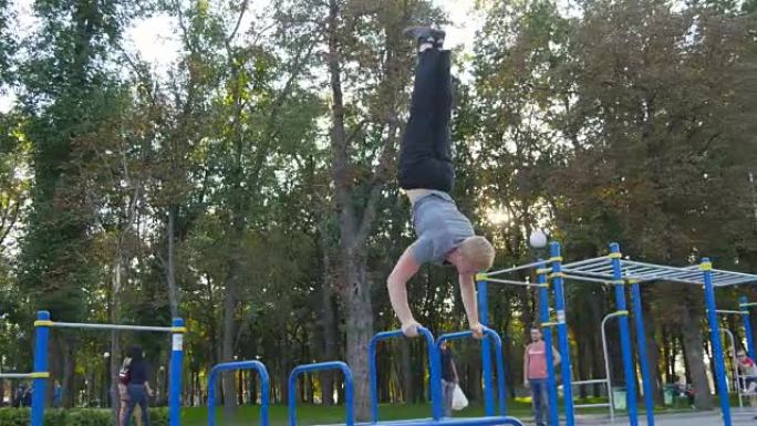 强壮的肌肉男人在公园倒立。适合肌肉发达的男性健身男子在室外单杠上做特技。运动员训练在室外肌肉处进行倒