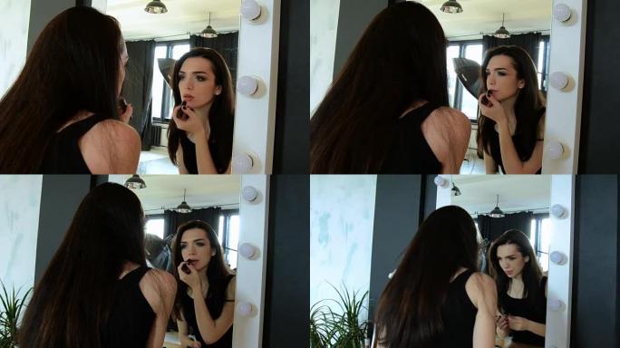 穿着黑色连衣裙的漂亮年轻黑发女人照镜子涂嘴唇