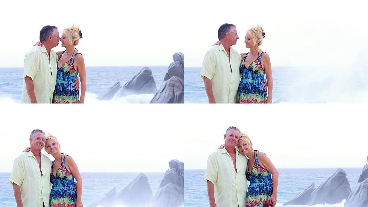一对年长的夫妇在海滩上互相亲吻，然后微笑，海浪拍打在他们身后的岩石上