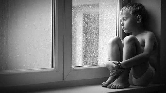 孤独的小男孩透过窗户玻璃看着雨滴，坐在窗台上