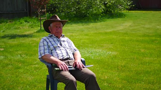 老人累了，坐在绿色草坪上戴帽子的椅子上。