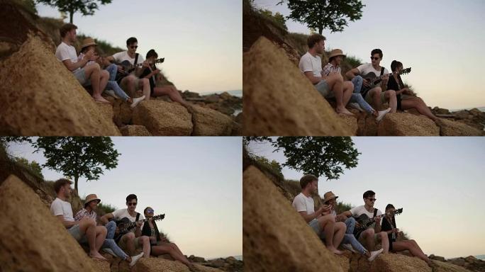 一群年轻开朗的人坐在海边的岩石上，弹吉他，唱歌和跳舞。慢动作镜头