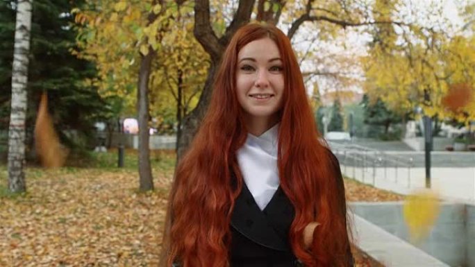 一个留着飘红头发的女孩站在公园落叶下。