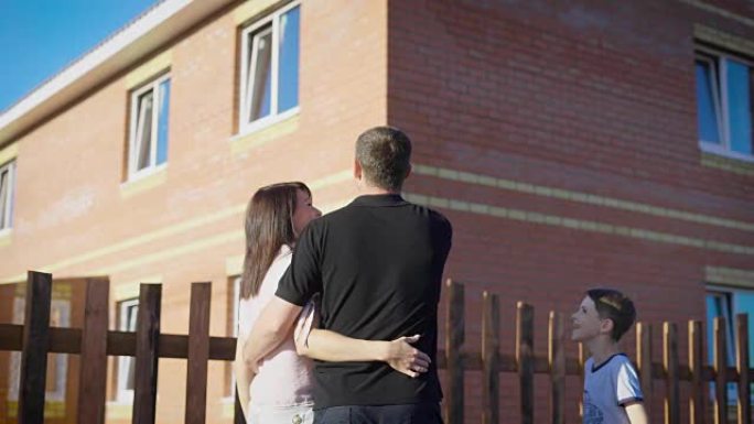 幸福的夫妇站在外面拥抱并看着新的围栏公寓。充满爱心的家庭在夏天阳光明媚的日子里展示男孩现代的房子。母