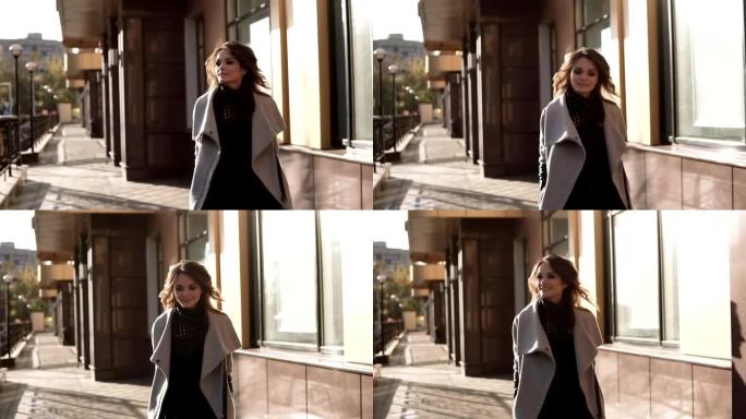 迷人的年轻女子穿着外套走过秋天的城市，微笑着。现代建筑背景下的可爱女孩。慢动作