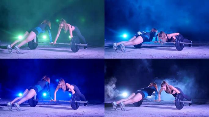 两个运动女孩成对地用杠铃做各种力量练习，俯卧撑，晚上，在淡淡的烟雾，雾中，在五彩缤纷的探照灯下，在一
