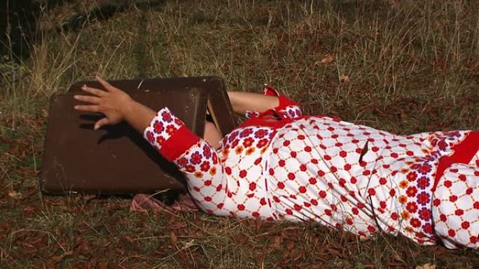 穿着白色圆点礼服的怪异女人躺在草地上，带着手提箱在大自然中