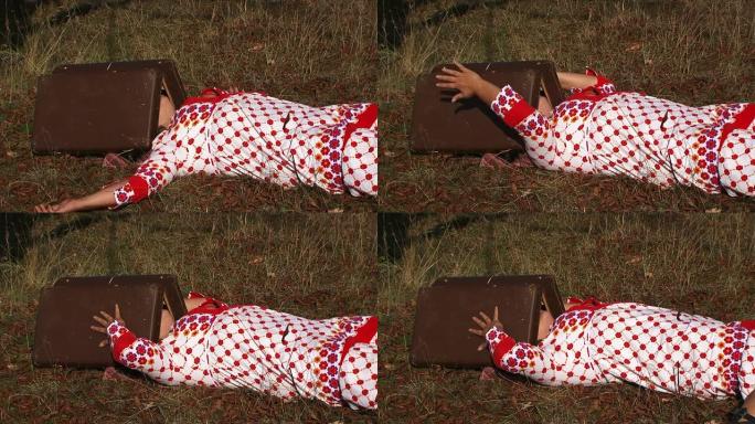 穿着白色圆点礼服的怪异女人躺在草地上，带着手提箱在大自然中