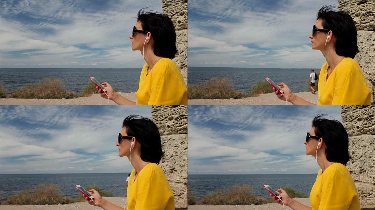 迷人的女人在智能手机上戴着耳机听音乐，背景是海洋和古城的废墟