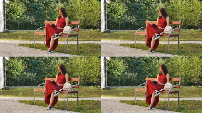 戴着眼镜和红色长裙的聪明女学生坐在公园的长凳上，在笔记本上写东西。