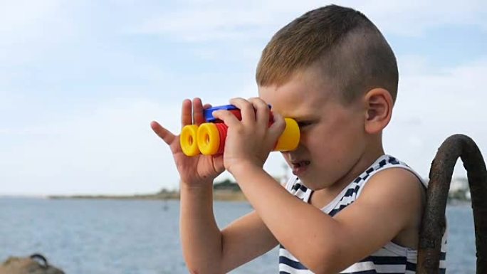 穿着条纹衬衫的漂亮小男孩在海边透过双筒望远镜看