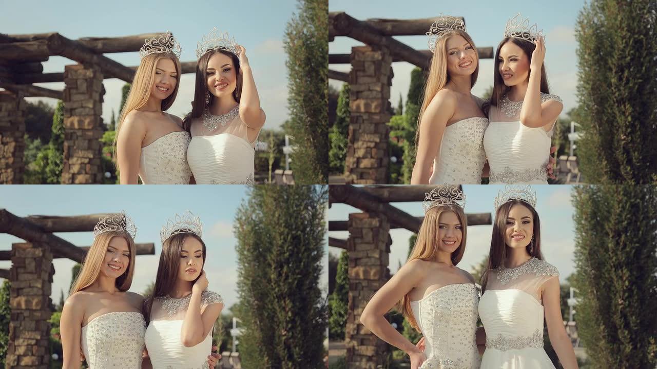 两个漂亮的女孩穿着优雅的白色连衣裙，头上的皇冠上绣有石头，在大自然的公园里摆姿势