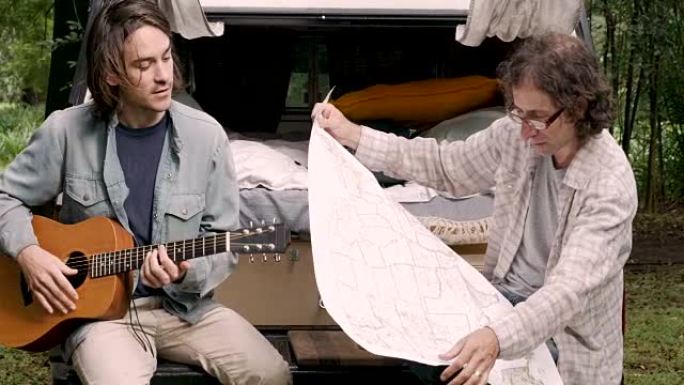 男子在露营时看着纸质地图，而他的男性朋友在弹吉他