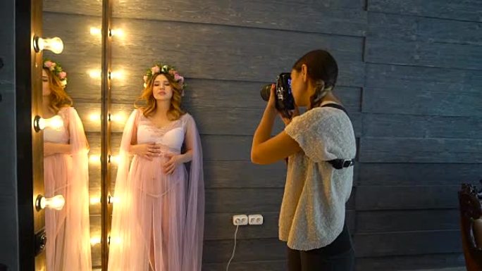 美丽的年轻怀孕照片模特在丁香peignoir为女摄影师摆姿势。