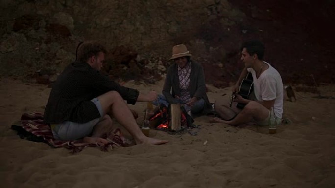 年轻朋友晚上坐在海边的火炉旁，烤香肠，喝啤酒，弹吉他。慢动作镜头