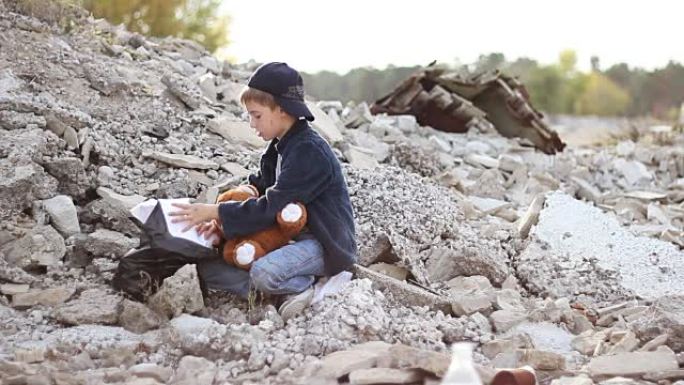 无家可归的孤儿男孩手里拿着一个柔软的玩具，在房屋废墟中寻找包装中的食物